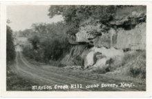 This real photo postcard looks north on K-4 Highway, two miles north of Dover, Kansas. The Highway crosses Mission Creek at the bottom of the hill.