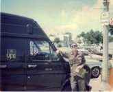Package car driver, Greg Hoots stands at the front of his 1979 Ford Econoline package car. The time card visible in the left shirt pocket and the fiber delivery-record clipboard date this photo 1982.
